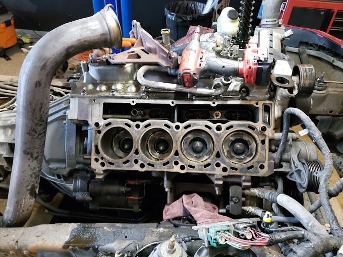 Partially disassembled engine block with visible cylinders and components placed on top. Tools and parts surround the setup, indicating engine repair or rebuild process, showcasing mechanical work involved in restoring vehicle performance in a workshop environment.