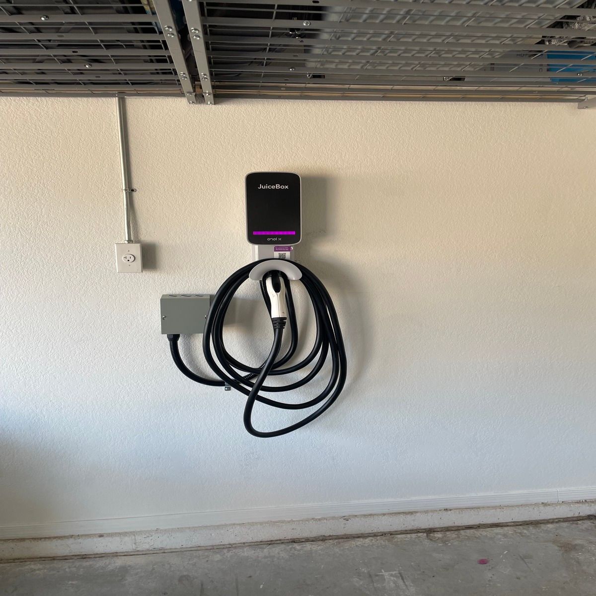 A JuiceBox electric vehicle charging station is mounted on a garage wall beneath overhead storage racks, with its long black cable neatly looped beside electrical conduit and a junction box.