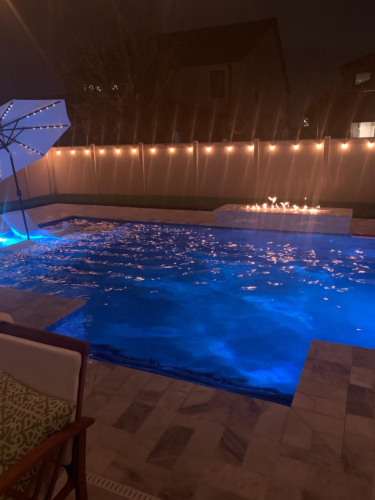 Residential swimming pool at night with bright blue underwater lighting, string lights, and a cozy outdoor patio setting.