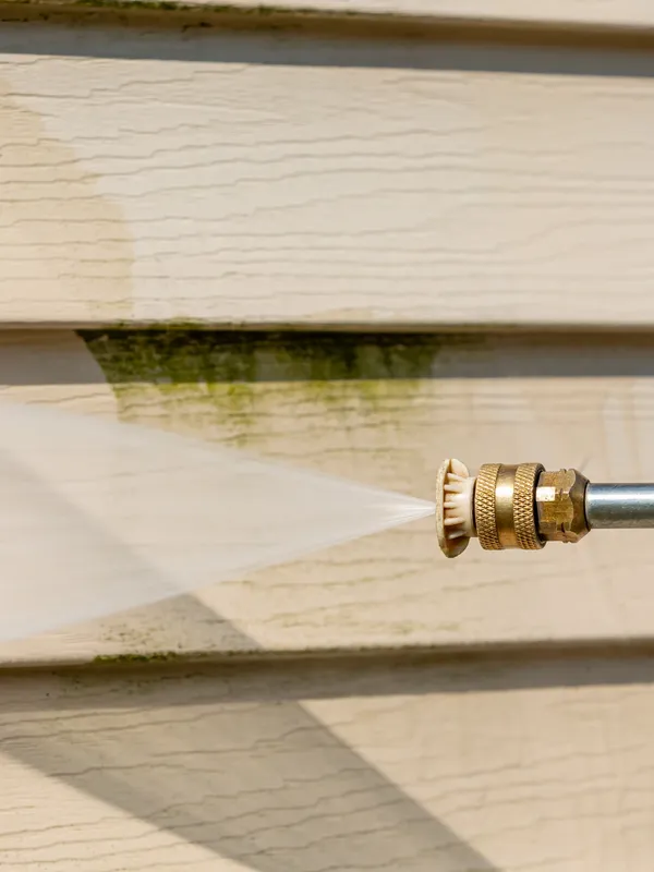 A close-up shot shows a pressure washer nozzle spraying water onto light-colored horizontal siding, effectively removing green algae and grime. The water spray creates a misty fan shape as it hits the surface, leaving a clean, wet path on the siding. The siding itself has a textured, wood-like grain pattern. Shadows from sunlight fall across the lower portion of the image.