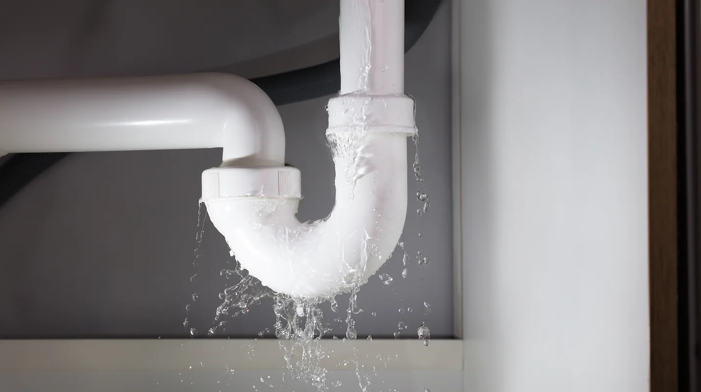 A close-up shot of a white PVC pipe under a sink with a noticeable leak. Water is actively spraying and dripping from a joint in the pipe, creating a splash effect against a dark gray background. The surrounding area appears to be cabinetry, with a hint of a wooden frame on the right edge of the image.