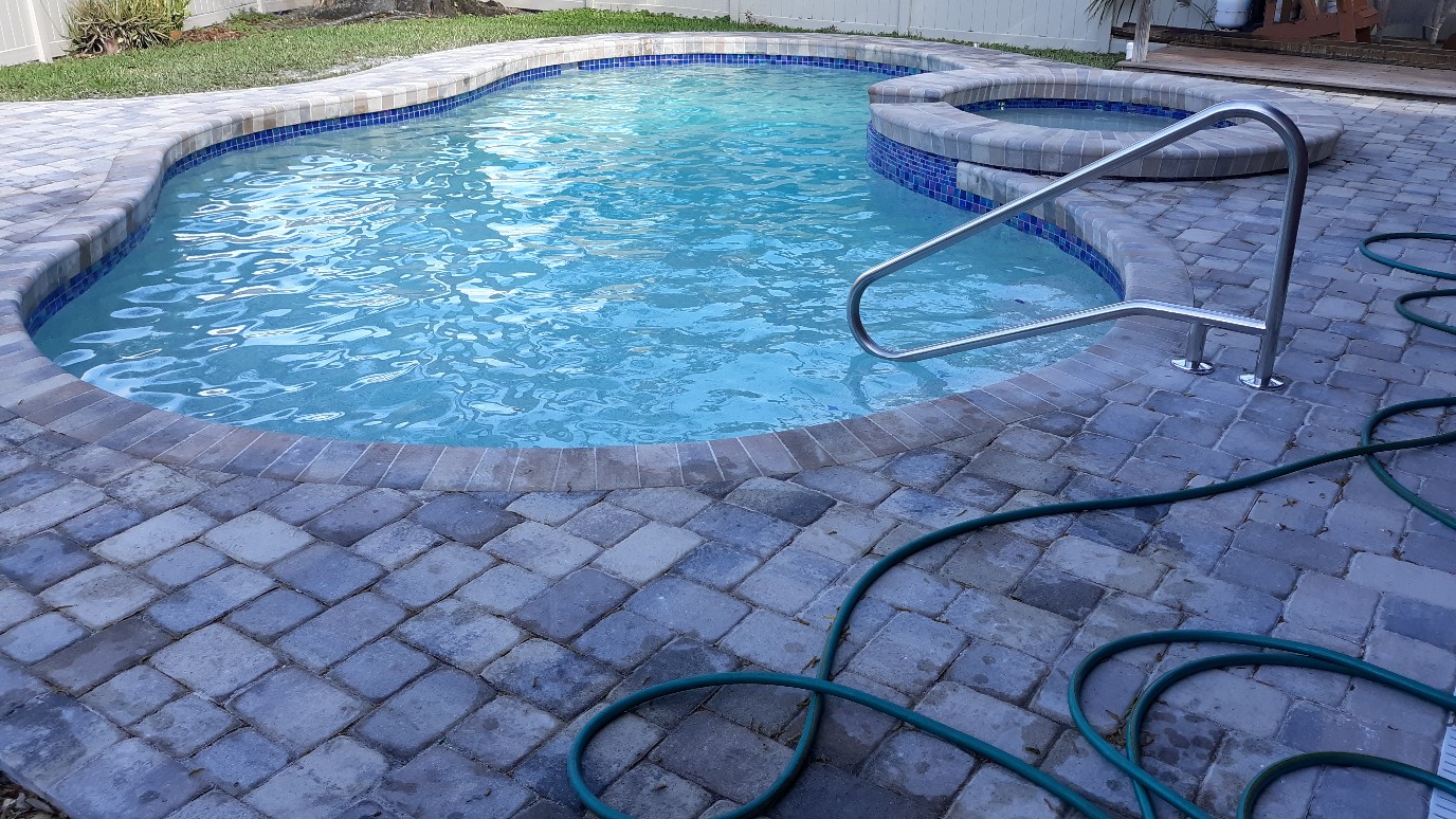 Freeform residential swimming pool with clear blue water, curved stone coping, brick paver decking, and a metal handrail.
