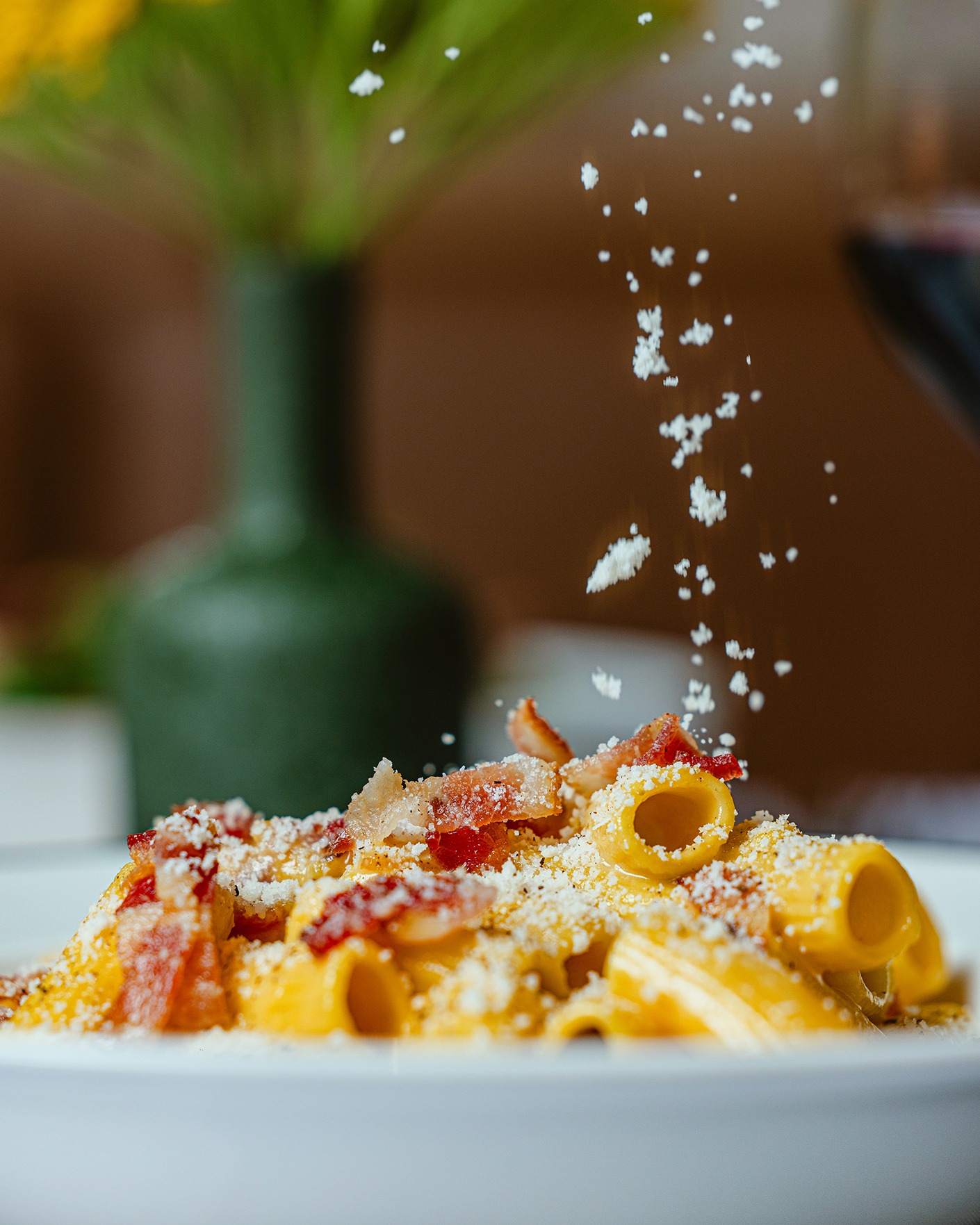 An action shot of finely grated cheese falling onto a plate of rigatoni carbonara topped with crispy pancetta.