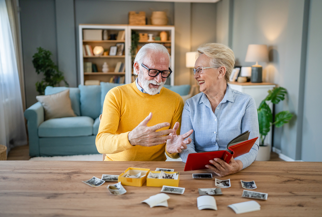 An elderly couple sits at a wooden table, smiling and reminiscing over old photos. The cozy room has a sofa, a lamp, and a bookshelf in the background.