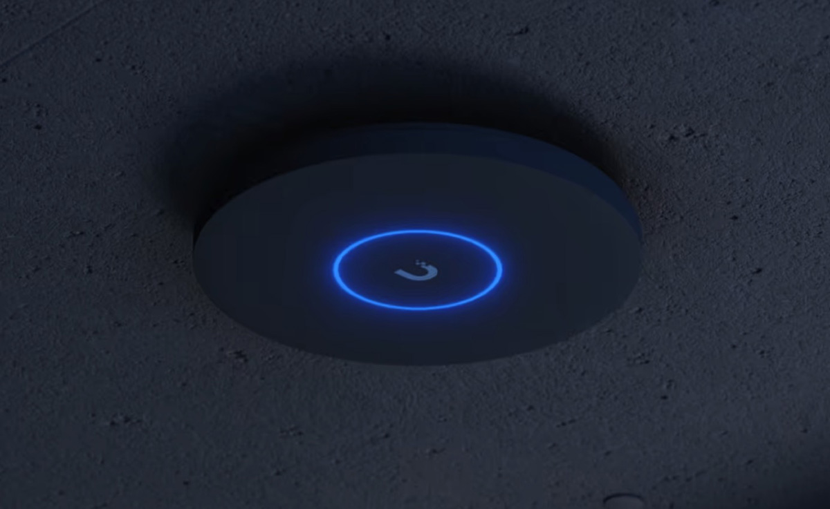 A close-up, angled view of a dark gray, circular device mounted on a textured, dark ceiling. The device has a bright blue LED light ring around a central symbol that resembles a lowercase 'u' with a dot above it, suggesting a modern technological product.