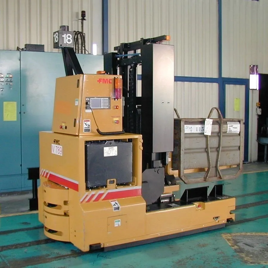 An automated guided vehicle (AGV) with a yellow and black body is parked in a warehouse or factory setting. The AGV has a tall mast and is holding a grey metal container. The container has labels on it. The AGV has "FMC V 789" printed on its side, and an "FMC" logo is visible on a control panel. The background shows industrial machinery, metal shelving, and a concrete floor. There is a sign with the number "18" displayed prominently on top of the AGV's housing. The overall scene suggests an operational industrial environment.