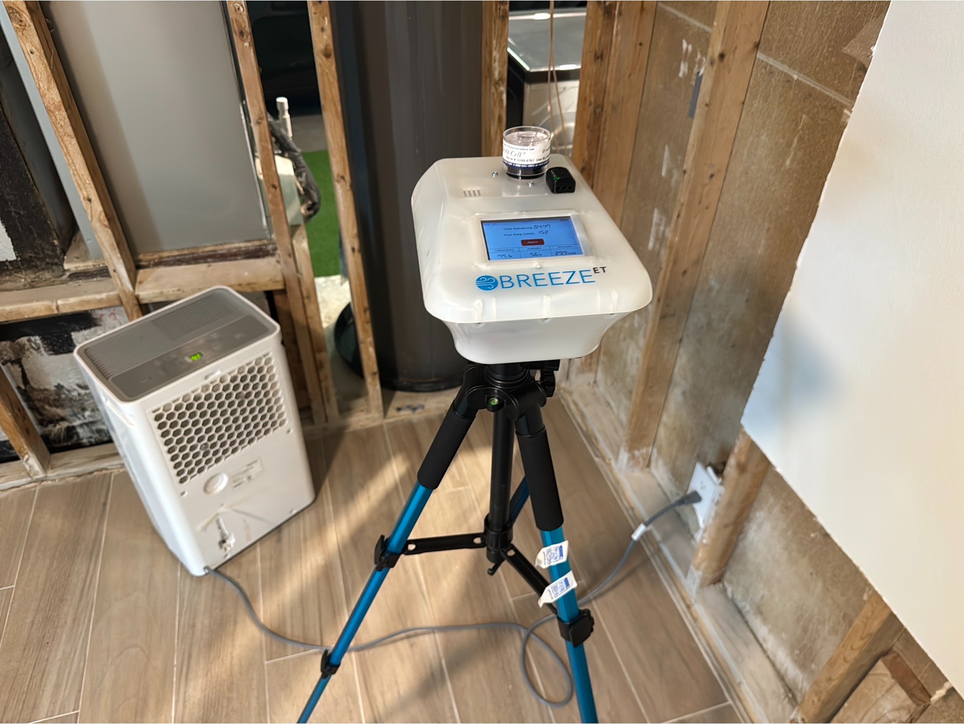 A Breeze ET device on a tripod is measuring air quality in a room with exposed wall studs. A portable air purifier is positioned nearby on the tiled floor.