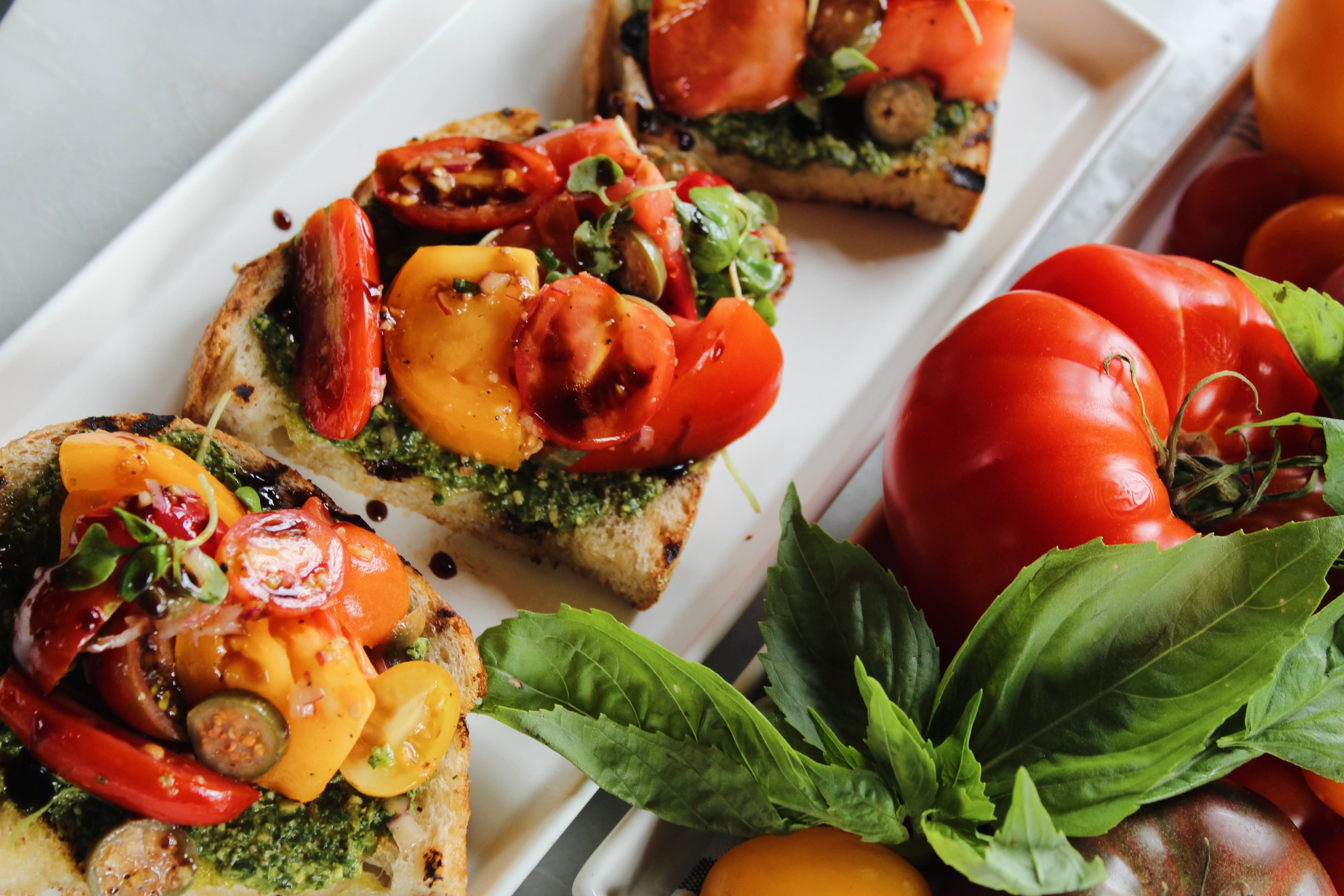 A platter of our Tomato Bruschetta — grilled rustic sourdough spread with basil pesto and heaped with jewel-toned heirloom tomatoes, finished with balsamic glaze.