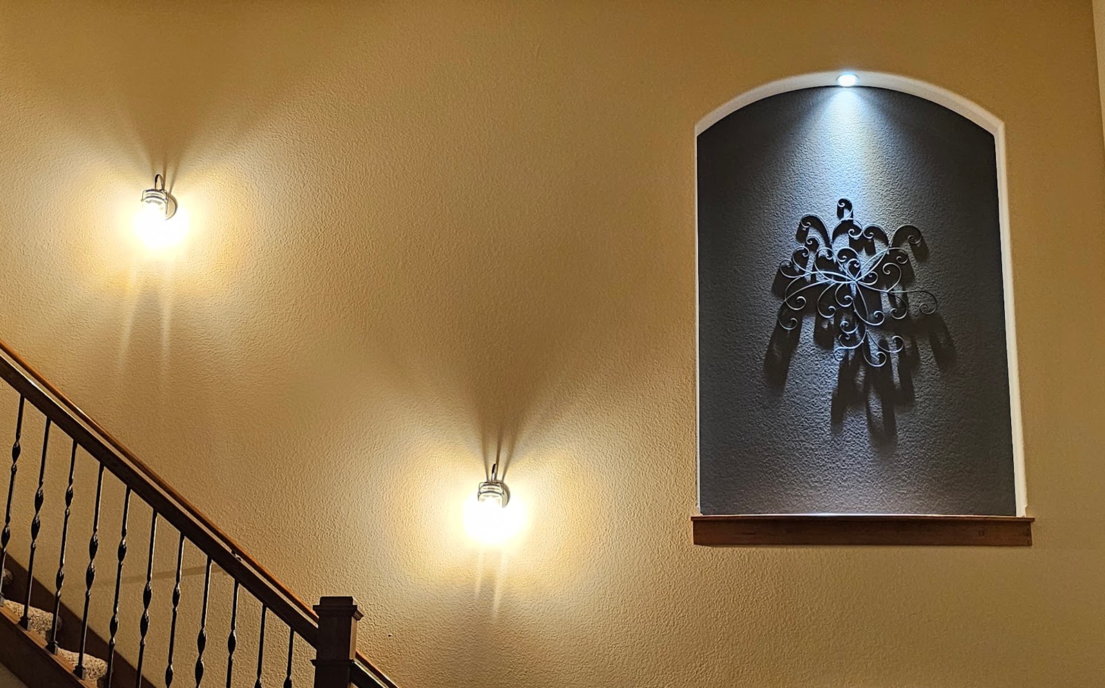 A dimly lit interior hallway shows a carpeted staircase with a wrought iron railing on the left. On the right, an arched niche in the wall displays a decorative metal wall art piece, possibly a sunburst or stylized bird, casting a shadow on the dark textured background. Two wall sconces with warm lighting illuminate the textured tan walls, casting soft shadows on the wall.