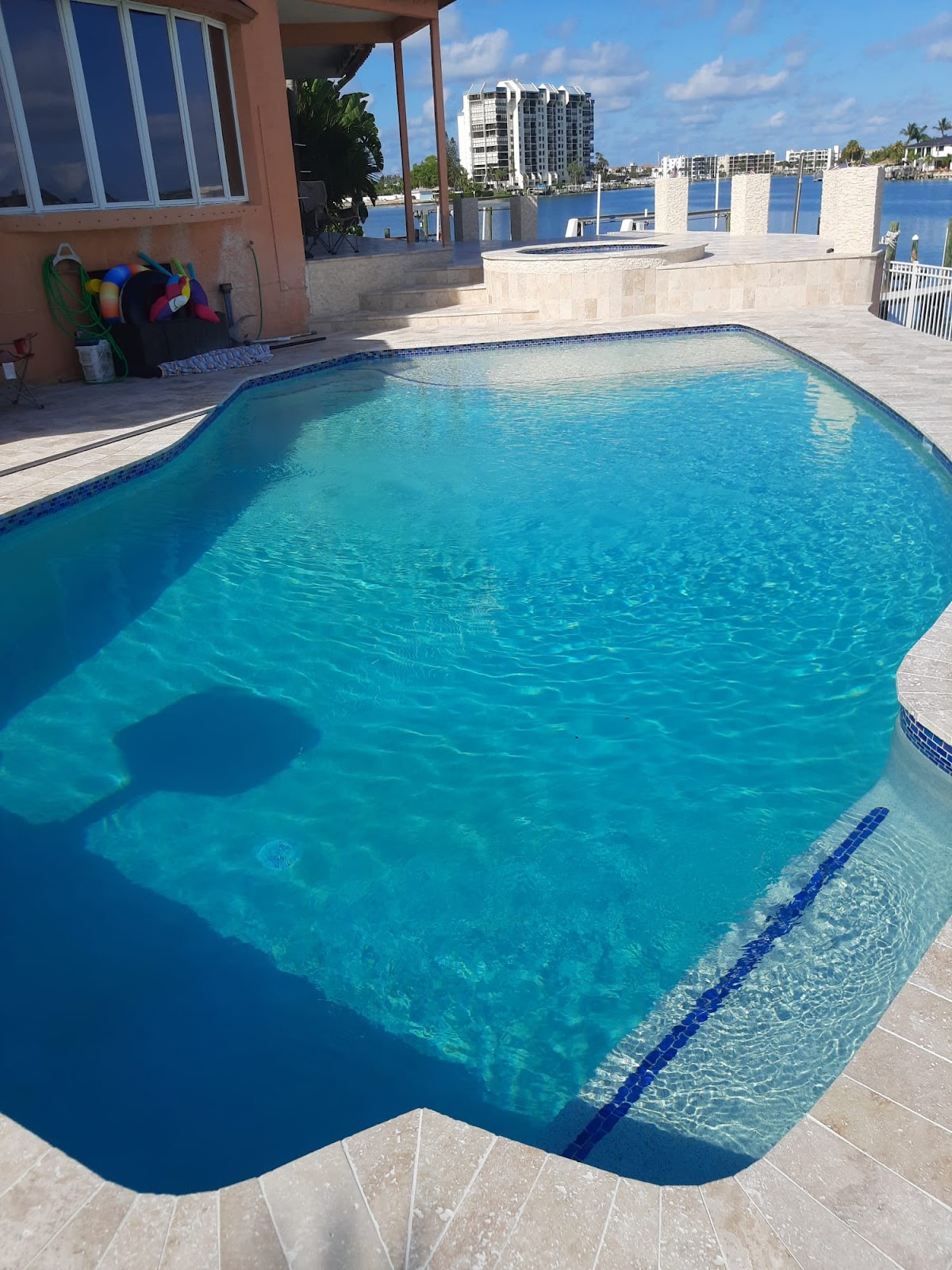 Waterfront residential swimming pool with bright turquoise water, light stone decking, and views of nearby buildings and the bay.