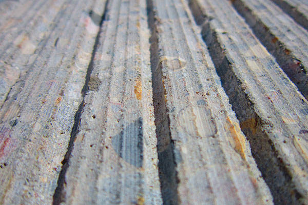 An extreme, low-angle macro close-up photograph showcasing the finish left by specialized diamond grinding. The surface displays perfectly parallel, deep corduroy-like longitudinal grooves. The view emphasizes the micro-texture of the exposed aggregate within the gray concrete, demonstrating how the specialized grinding process creates a high-friction, uniform surface, likely Next Generation Concrete Surface (NGCS)