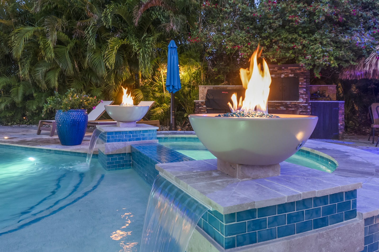 Luxury swimming pool and spa at night featuring decorative fire bowls, colorful pool lighting, and lush tropical landscaping.