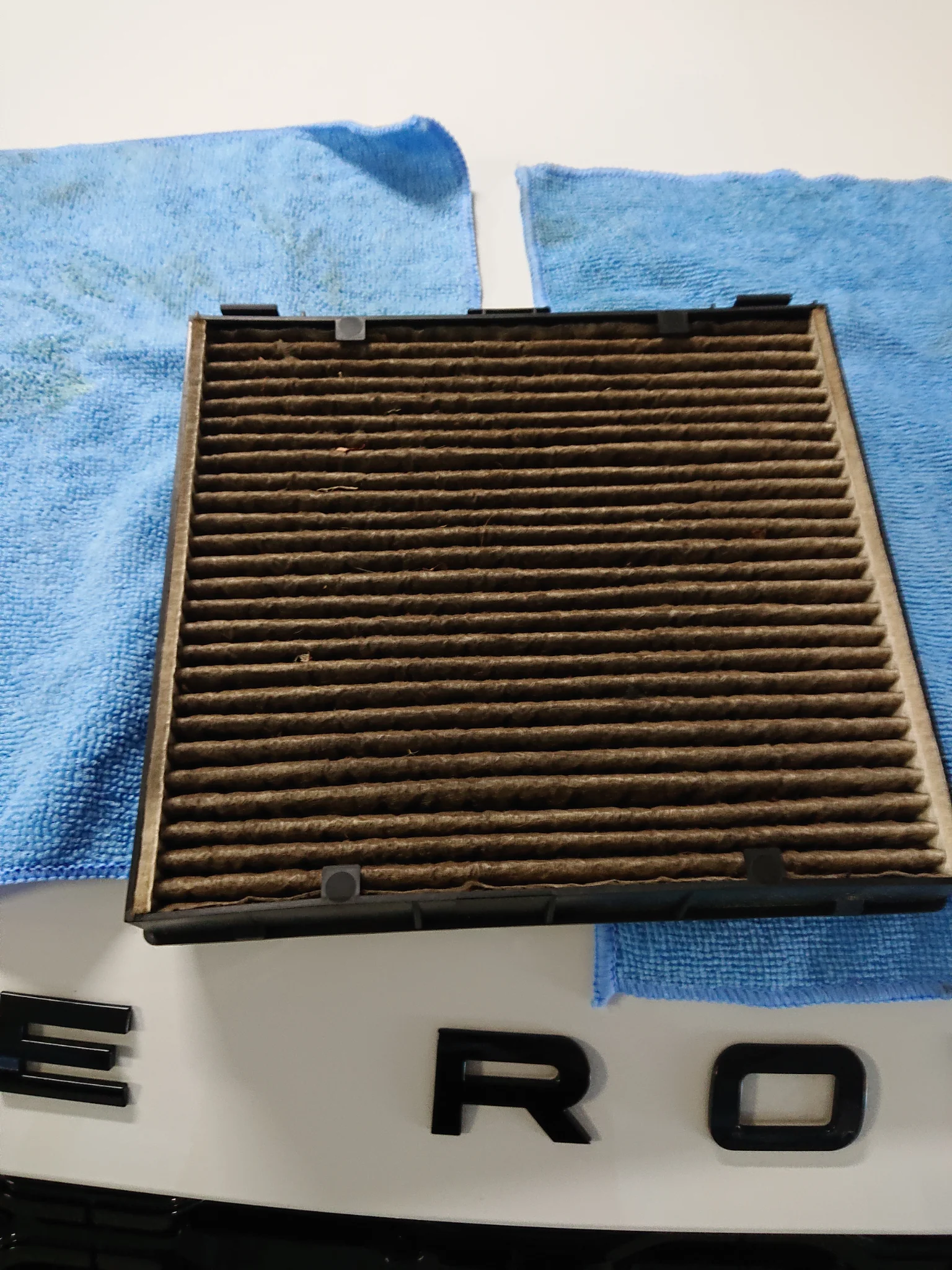 A dirty cabin air filter sits on top of a white surface with black lettering. Two blue microfiber cloths are placed underneath and to the sides of the filter.