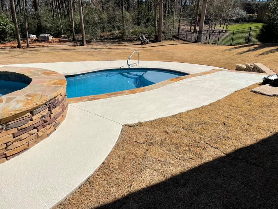 A newly constructed concrete pool deck surrounds a bright blue swimming pool. To the left, a circular spa feature is clad in stacked stone. A pathway of smooth, light-colored concrete leads from the spa area towards the pool. The surrounding area is covered in dry, brown grass with some areas of newly laid sod. In the background, mature trees line a black metal fence, and some construction materials are visible on the left side of the frame. The scene appears to be a backyard renovation project.