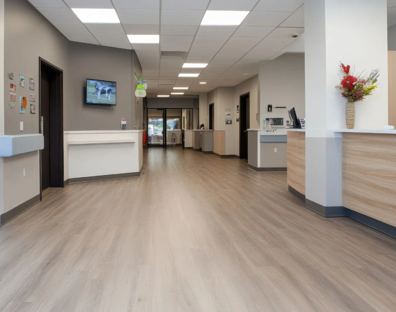 A clean, wide healthcare lobby and corridor featuring light wood-look vinyl flooring, neutral gray walls, recessed LED ceiling lighting, a reception desk with wood paneling, and wall-mounted monitors.