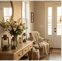A bright and airy entryway features a wooden console table adorned with a large vase of flowers, two antique lanterns, and a small jar. To the right, a comfortable armchair draped with a cozy blanket sits near a white door with glass panes. A round mirror hangs on the wall above the console table, reflecting the natural light. The overall aesthetic is warm, inviting, and hints at a rustic or farmhouse style.