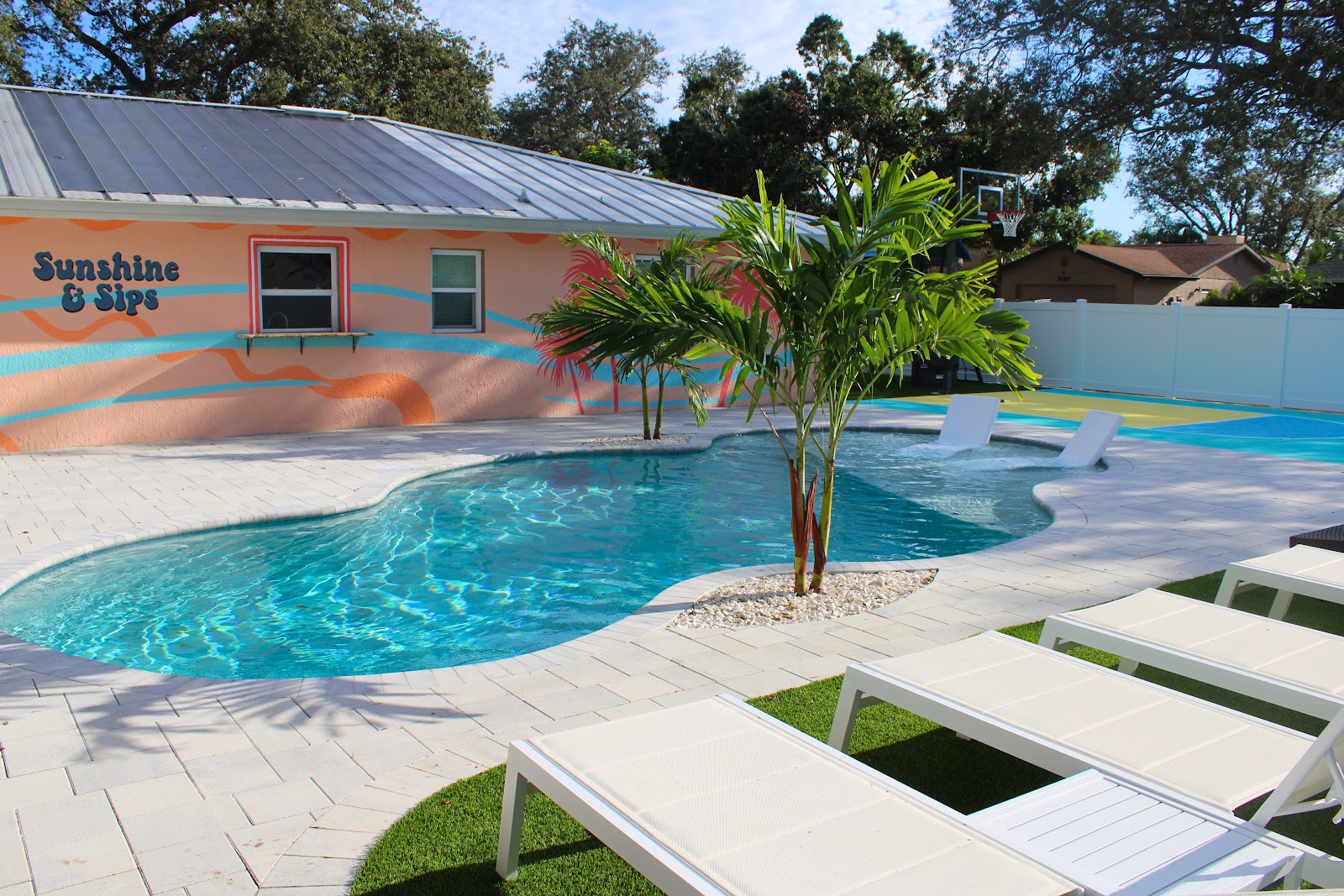 Bright backyard pool area with curved swimming pool, light paver decking, palm trees, lounge chairs, and a colorful tropical-style home.