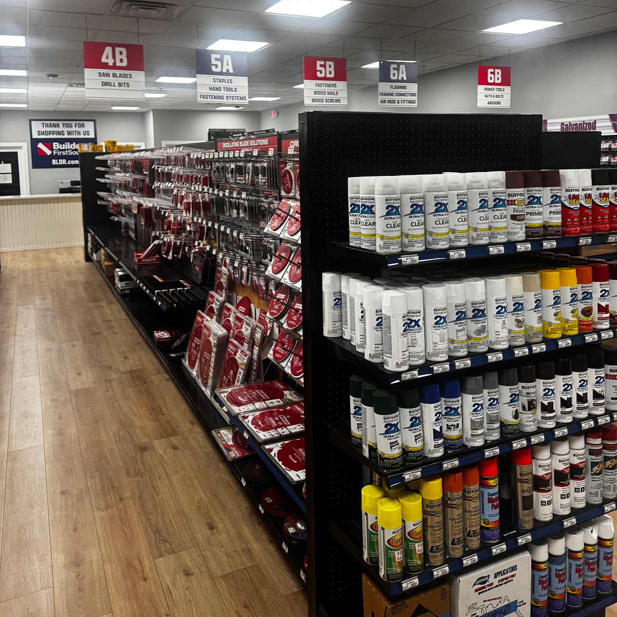 Wide view of a Builders FirstSource hardware department with black shelving and pegboard displays. Shelves on the left hold packaged Schlage locksets, while center and right displays contain door hardware, gate hardware, electrical items and bright yellow tie-down straps. Overhead signs label sections for locksets, door hardware, electrical, sandpaper, drywall tools and accessories, and other hardware.
