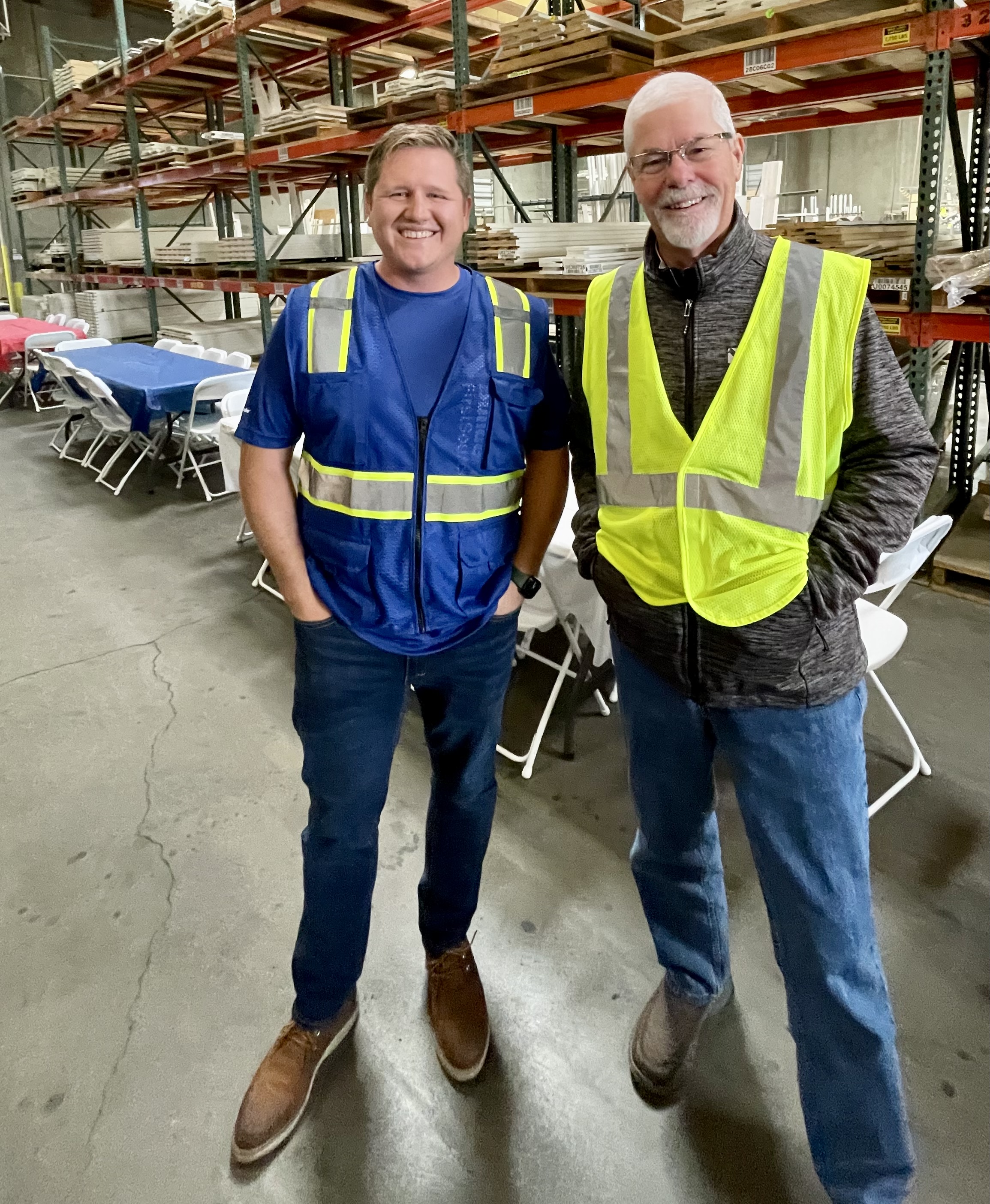 Two men wearing safety vests and casual clothing are standing in a warehouse, smiling at the camera. The man on the left is wearing a blue vest, and the man on the right is wearing a yellow vest.