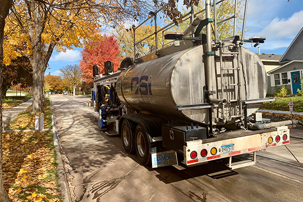 A rear perspective photograph of a large, complex truck-mounted specialized grinding machine. The rig is staged curbside on an urban or residential street lined with mature trees displaying vibrant yellow and orange autumn foliage. The vehicle is a massive, multi-component piece of heavy construction equipment with complex hydraulic lines and a large vacuum slurry management system, signaling active work in a tight environment.