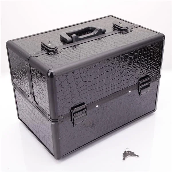 A closed black makeup case with a textured crocodile-like pattern rests on a white surface. The case has a black handle on top, silver clasps on the front and sides, and black metal trim. A small silver key lies on the surface to the right of the case. The background is a plain white.
