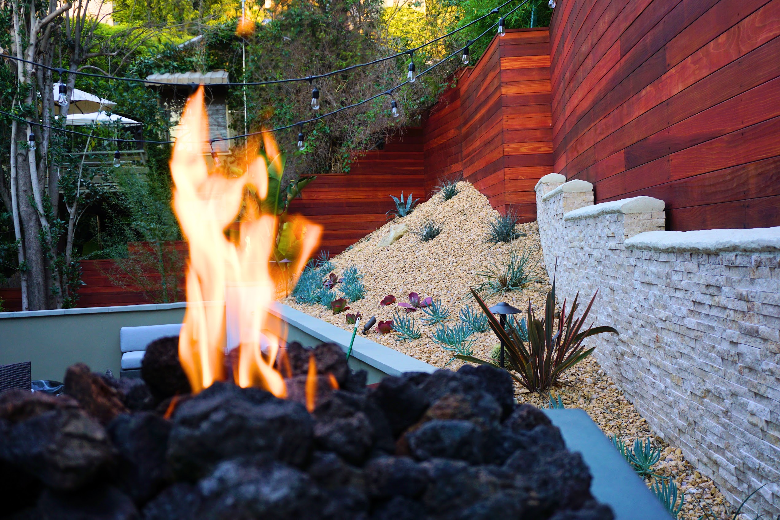 Flames rise from a black rock fire pit beside a landscaped succulent garden, red wood fence, string lights, and white stone wall.