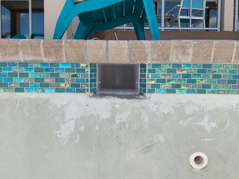 Close-up of a swimming pool wall with decorative blue and green tile, coping, and a built-in water feature opening.