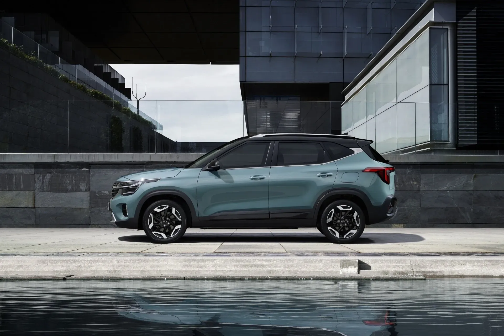 A teal Kia Seltos SUV is parked on a paved area next to a body of water, with a modern building in the background. The building features dark-colored stone and glass facade with a stepped architectural design.