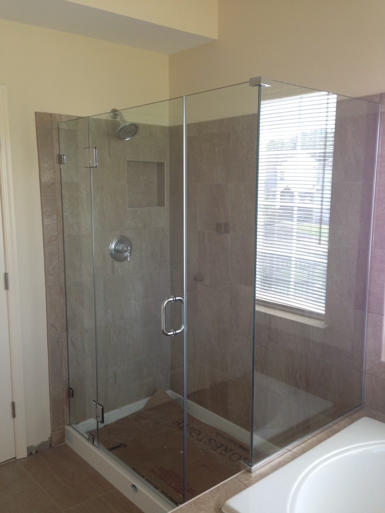 A modern glass shower enclosure with a brushed nickel shower head and handle is installed in a bathroom with tan tile walls and a window with blinds. The floor of the shower enclosure is covered with cardboard, and a white bathtub is visible to the right.
