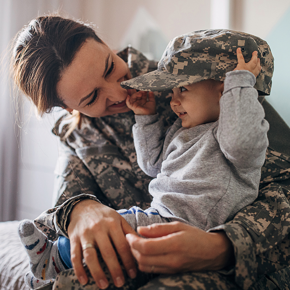 A woman wearing a camouflage uniform holding a young child wearing a camouflage hat.