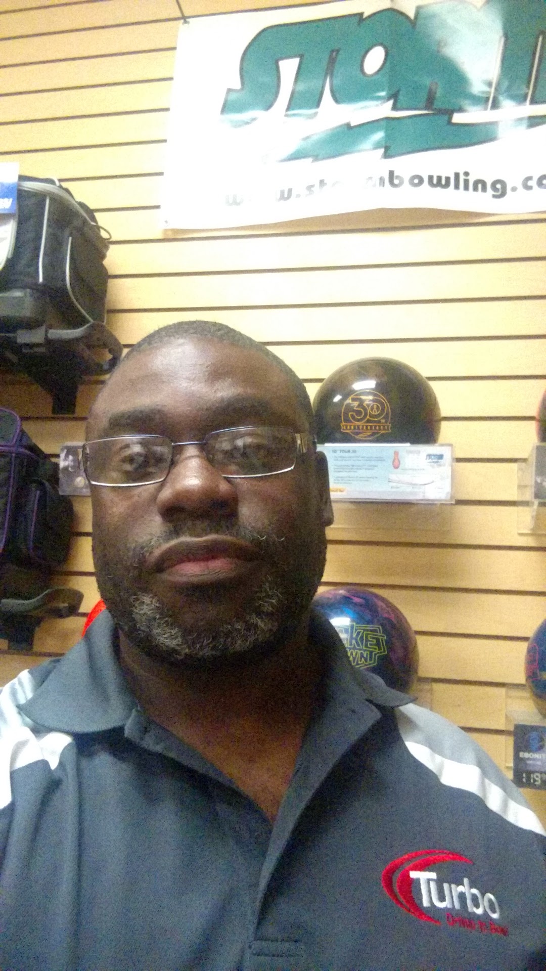A man with glasses and a beard takes a selfie in what appears to be a bowling alley pro shop. He is wearing a dark grey polo shirt with white stripes on the sleeves and a "Turbo" logo on the chest. Behind him, bowling balls and bags are displayed on shelves. A large banner with "Storm" logo is visible at the top.