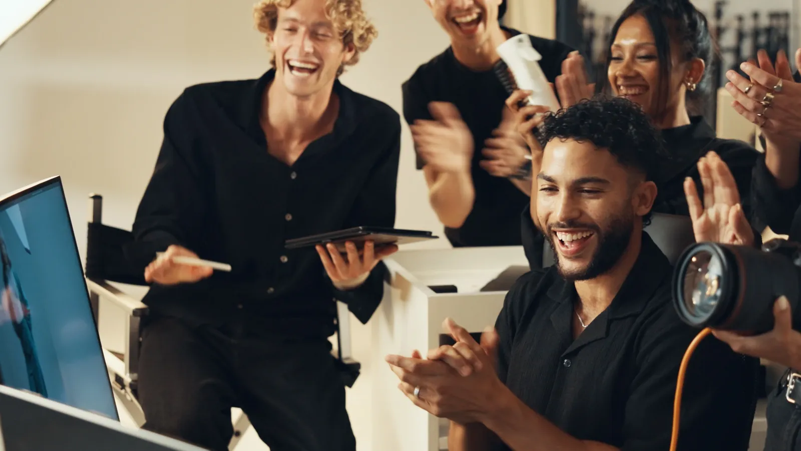 A diverse group of people are gathered, all smiling and appearing to be in good spirits. In the foreground, a man with curly dark hair is clapping, wearing a black short-sleeved shirt and a silver necklace. To his left, another man with blonde curly hair and a black button-up shirt is holding a tablet and a stylus, also laughing heartily. Behind them, several other individuals are visible, some clapping and one holding a professional camera. The scene suggests a collaborative and celebratory atmosphere, possibly related to a creative project or a successful event.