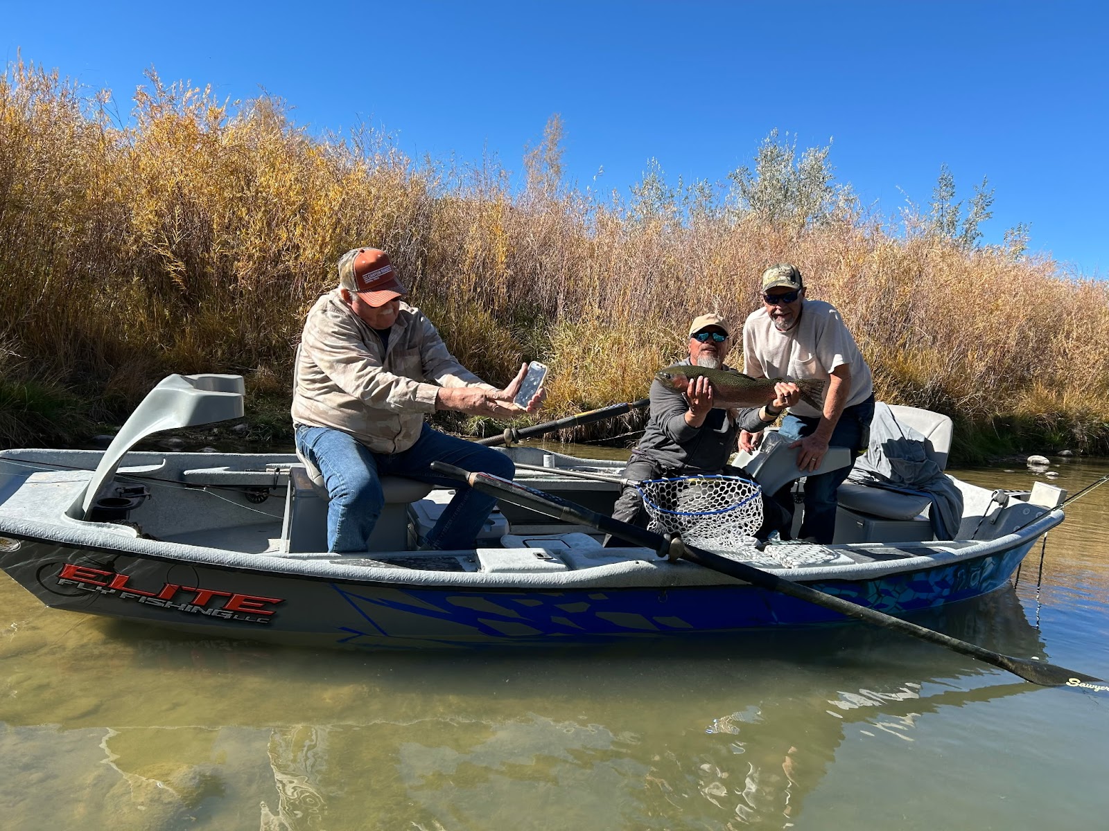 Three men in a boat on a sunny day, holding a large fish. The man on the left is holding up a phone, possibly taking a picture or video. The boat is branded 'ELITE FLY FISHING LLC'. The background shows a clear blue sky and dry, golden-colored reeds along the riverbank.