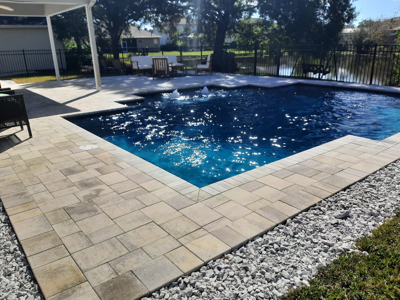 Residential backyard swimming pool with deep blue water, light stone paver decking, and a modern outdoor living area.