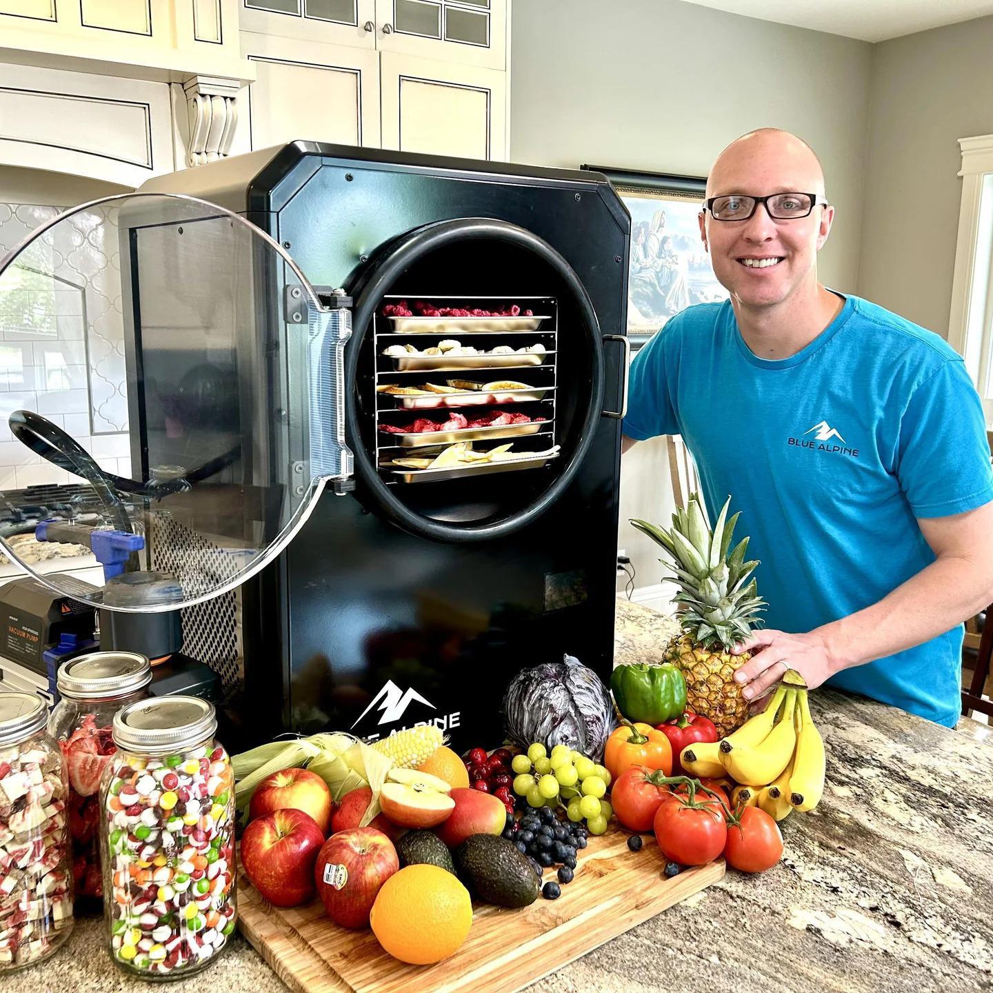 This image captures a moment in a well-lit, spacious kitchen where an individual with their face obscured is presenting a variety of fresh fruits and candies. The person is standing next to a large, black BLUE ALPINE dehydrator filled with slices of fruits. The countertop is adorned with an array of colorful fruits including bananas, apples, grapes, and a pineapple, as well as jars filled with assorted candies.