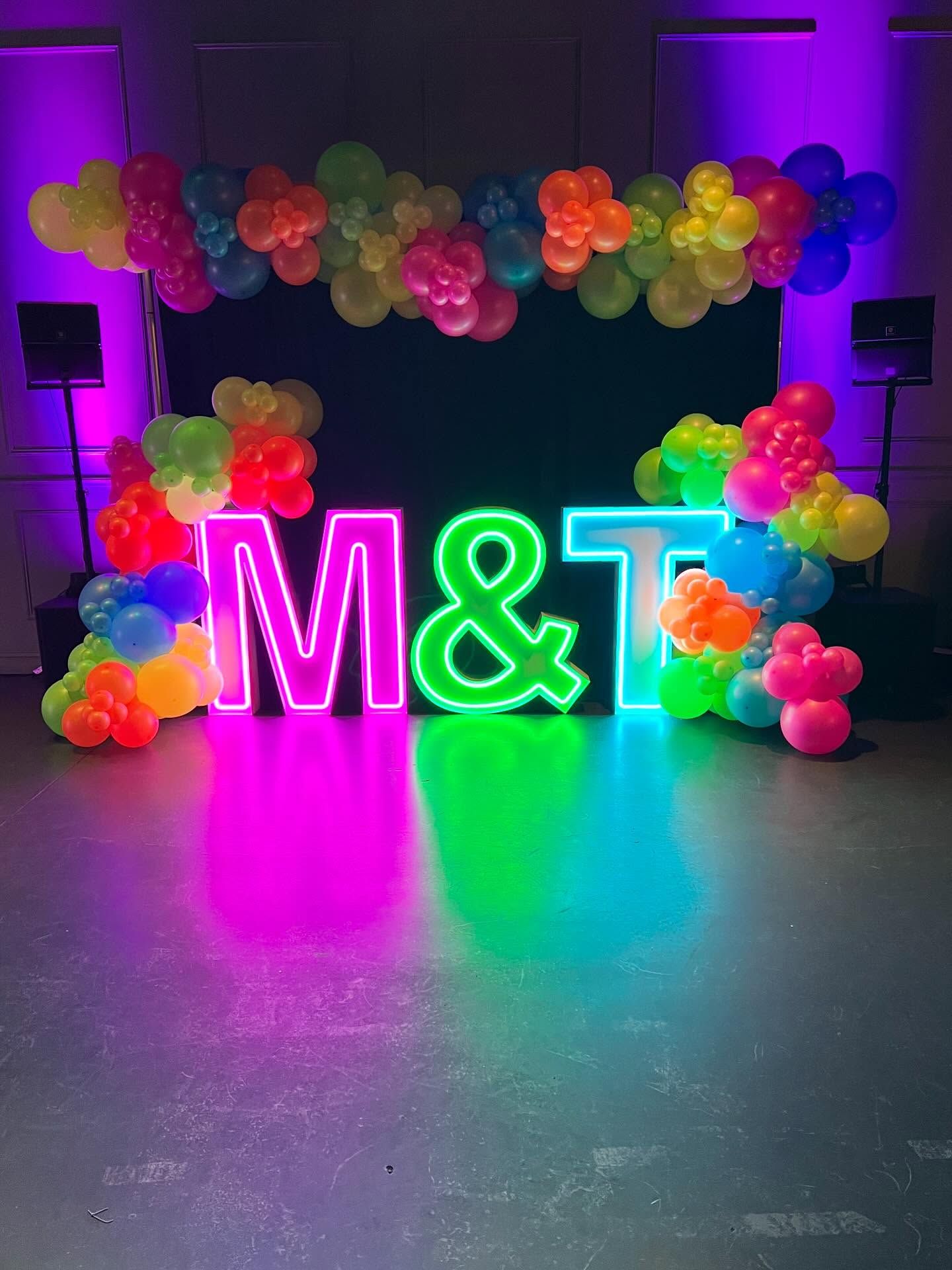 Vibrant event backdrop featuring neon M and T letters with glowing ampersand, surrounded by rainbow balloon garlands, purple uplighting, and dark stage setting, creating a lively celebratory photo moment scene.