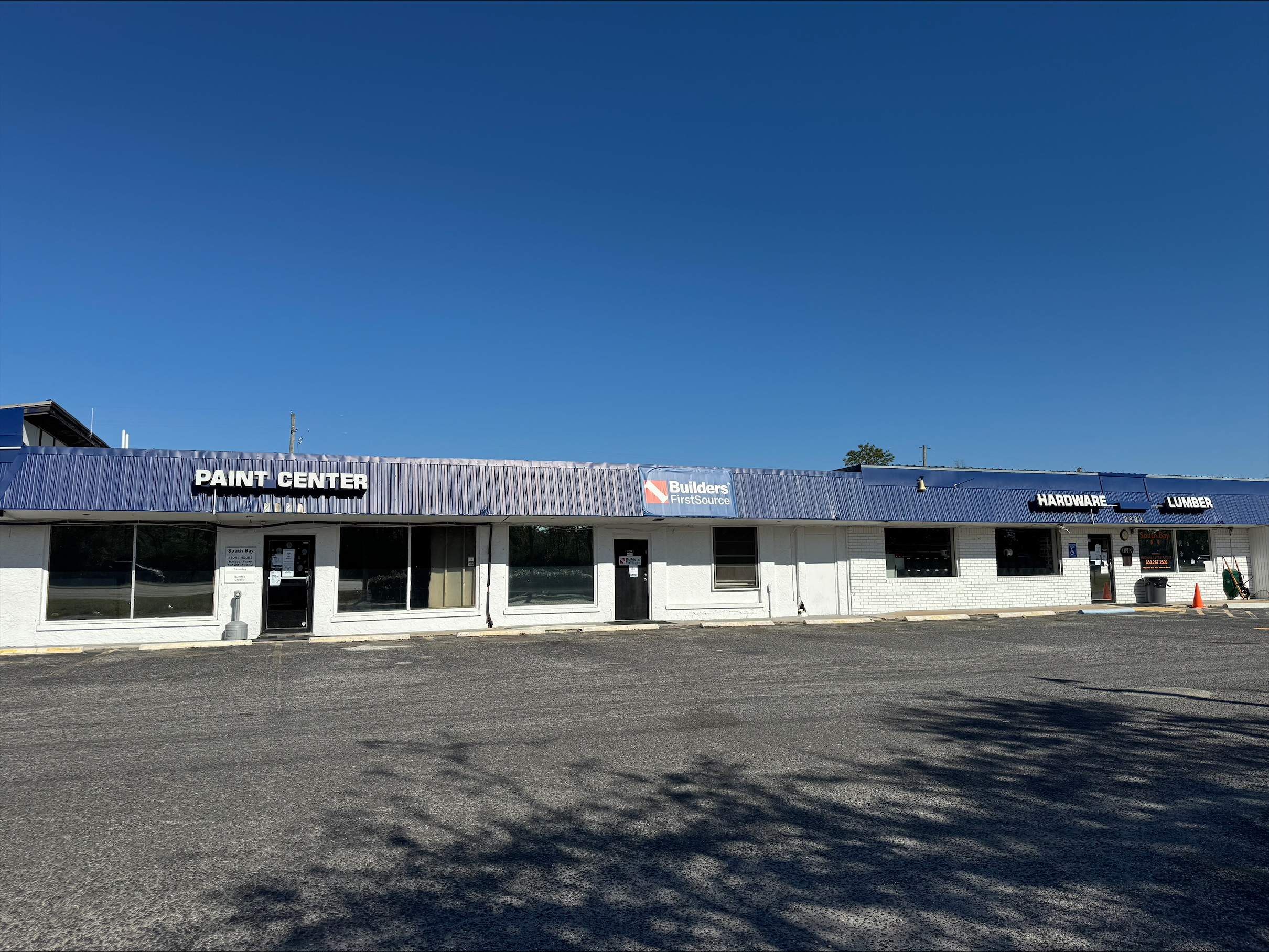 Front exterior view of the Builders FirstSource Lumber Yard in Santa Rosa Beach, Florida, showcasing the building with a blue awning labeled 'Paint Center,' 'Hardware,' and 'Lumber.