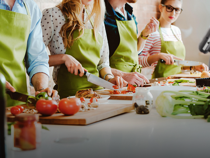 A group of four people, dressed in aprons, are engaged in a cooking class. They are actively chopping vegetables on wooden cutting boards, with fresh ingredients like tomatoes, bell peppers, and leafy greens spread around them. The scene suggests a collaborative and hands-on culinary experience.