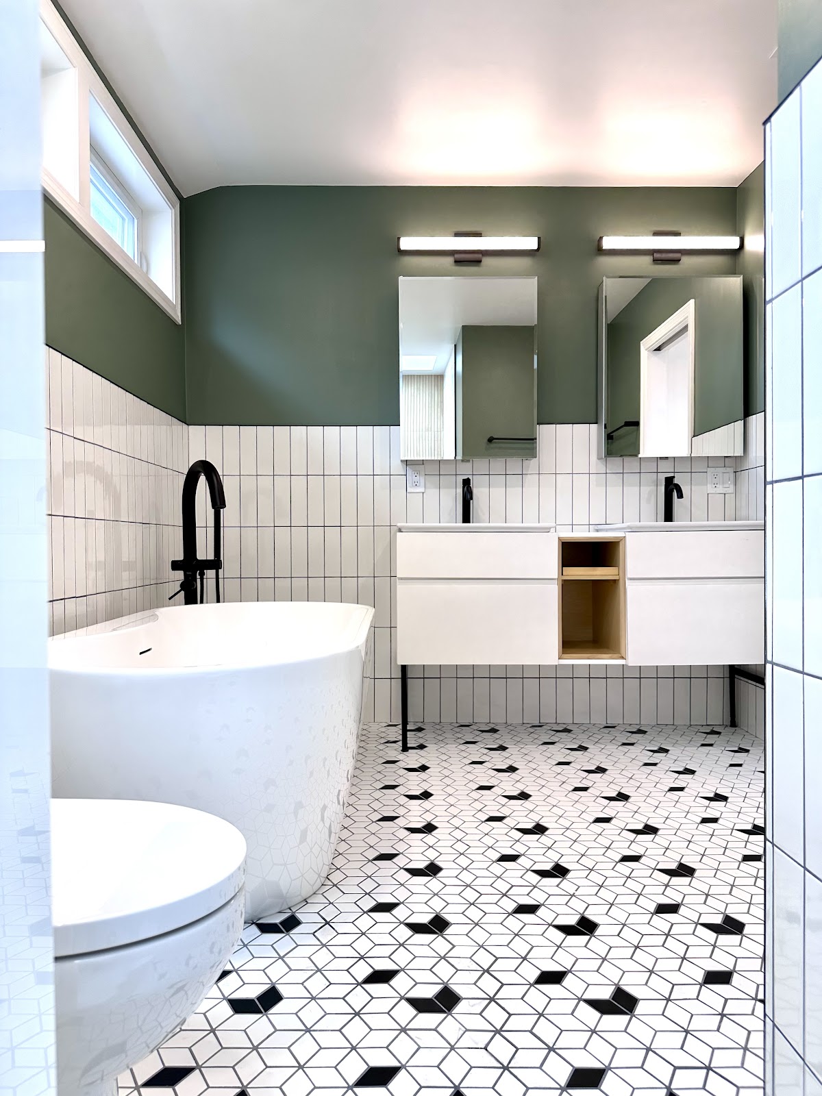 A modern bathroom featuring a freestanding white bathtub, a toilet, a white double vanity with black faucets, and two square mirrors above the vanity. The walls are tiled with white subway tiles up to a green painted section. The floor is decorated with a geometric pattern of black and white tiles. Linear light fixtures are mounted above the mirrors.