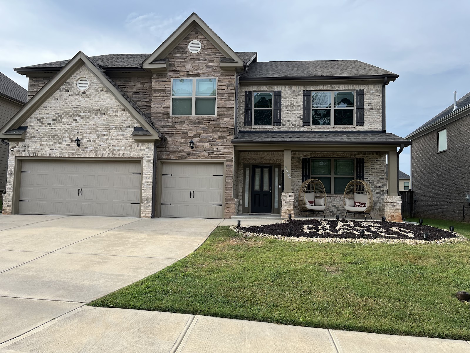 A two-story house with a three-car garage and a welcoming front porch with two hanging chairs. The house features a mix of beige brick and stone facade, dark shutters on the windows, and a gray roof. The landscaping includes a green lawn, a mulched flower bed with decorative rocks spelling out "RELAX", and several small outdoor lights. The sky is overcast.