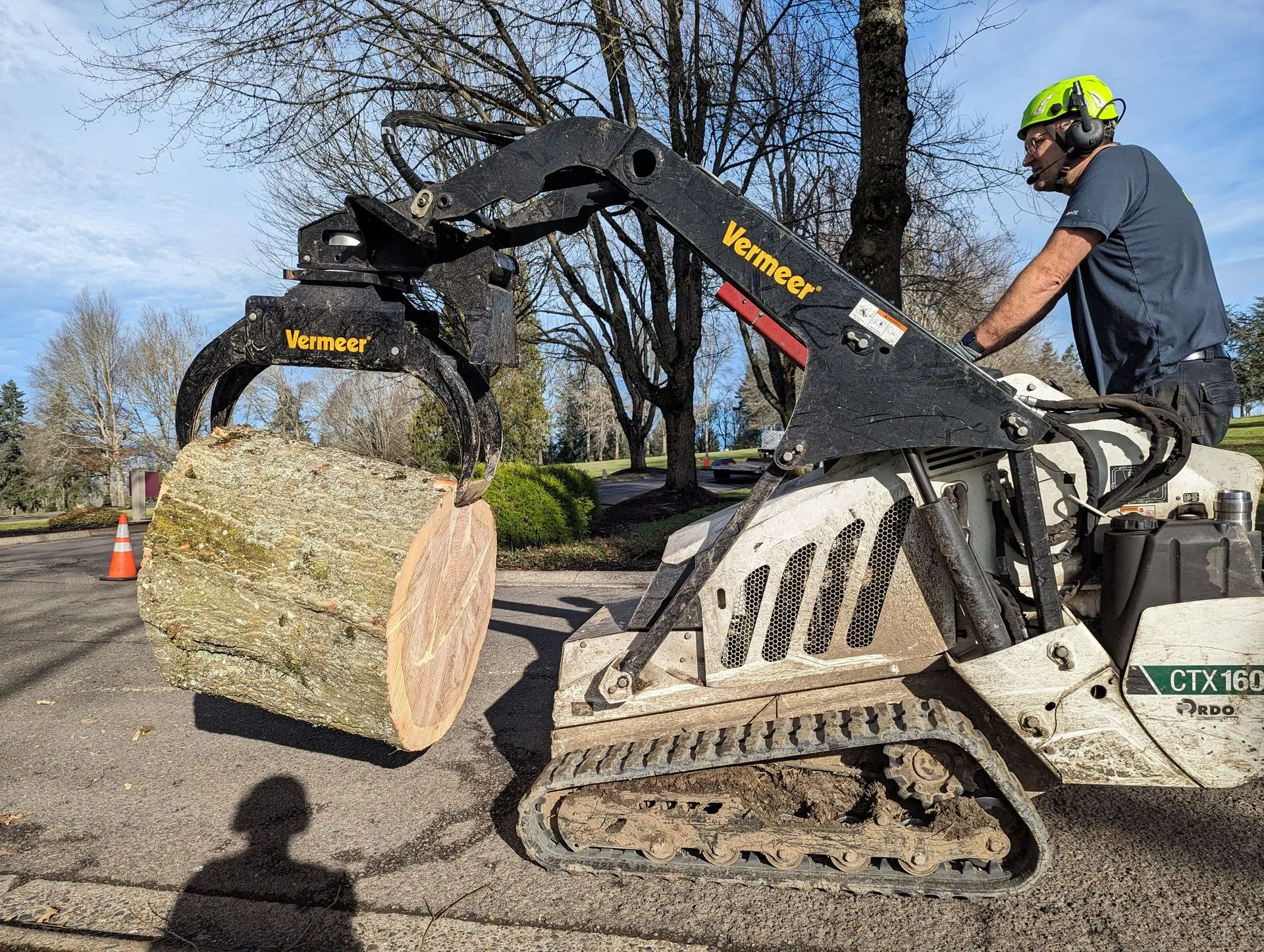 A compact track loader lifts a heavy tree section using a grapple attachment. The image highlights a 