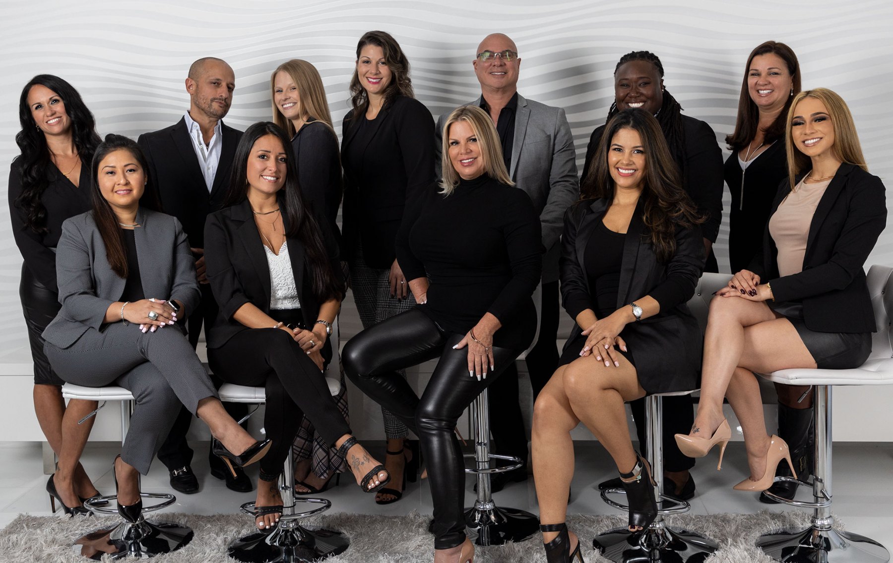 A group of eleven diverse professionals, comprised of six women and five men, are posing for a studio photograph. They are all dressed in formal business attire, predominantly black and gray suits and blazers, with some individuals opting for black tops. The setting is a clean, modern studio with a white, wavy textured wall as the backdrop. The group is arranged in two rows, with some standing and others seated on chrome-legged stools. They are all looking towards the camera with confident smiles, suggesting a cohesive and professional team.