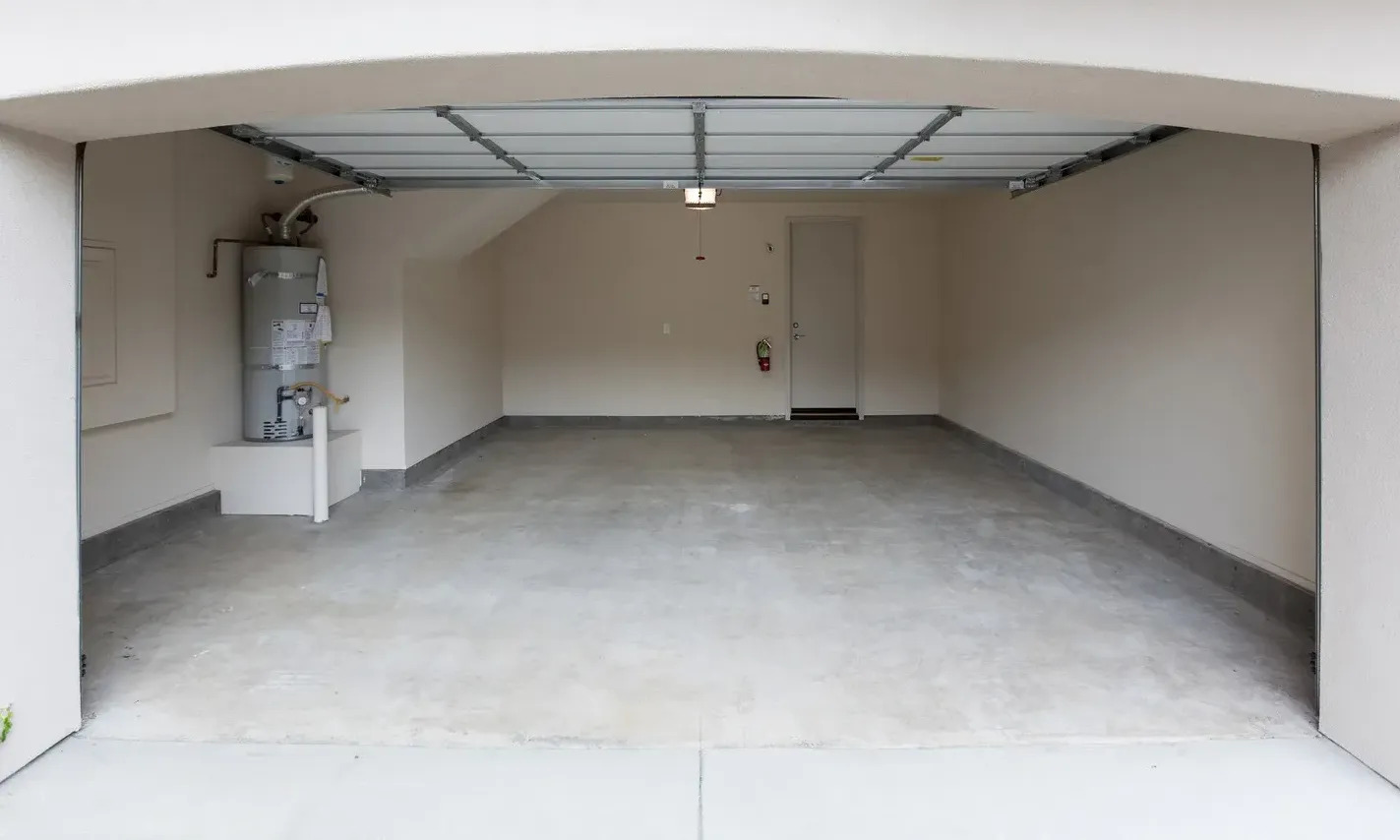 An empty, well-lit garage features a concrete floor, a partially open metal garage door, and a water heater. The space is clean and ready for use.