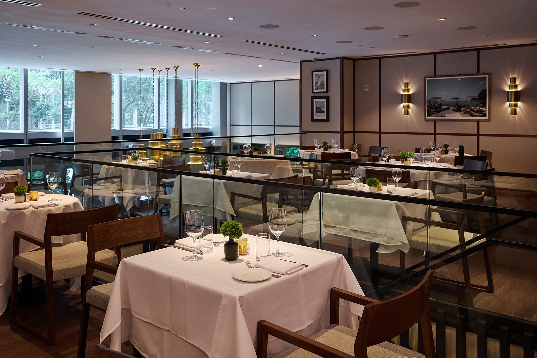An elegant view of a second-level dining area featuring glass railings, mid-century modern wooden chairs, and minimalist table settings overlooking the main floor.