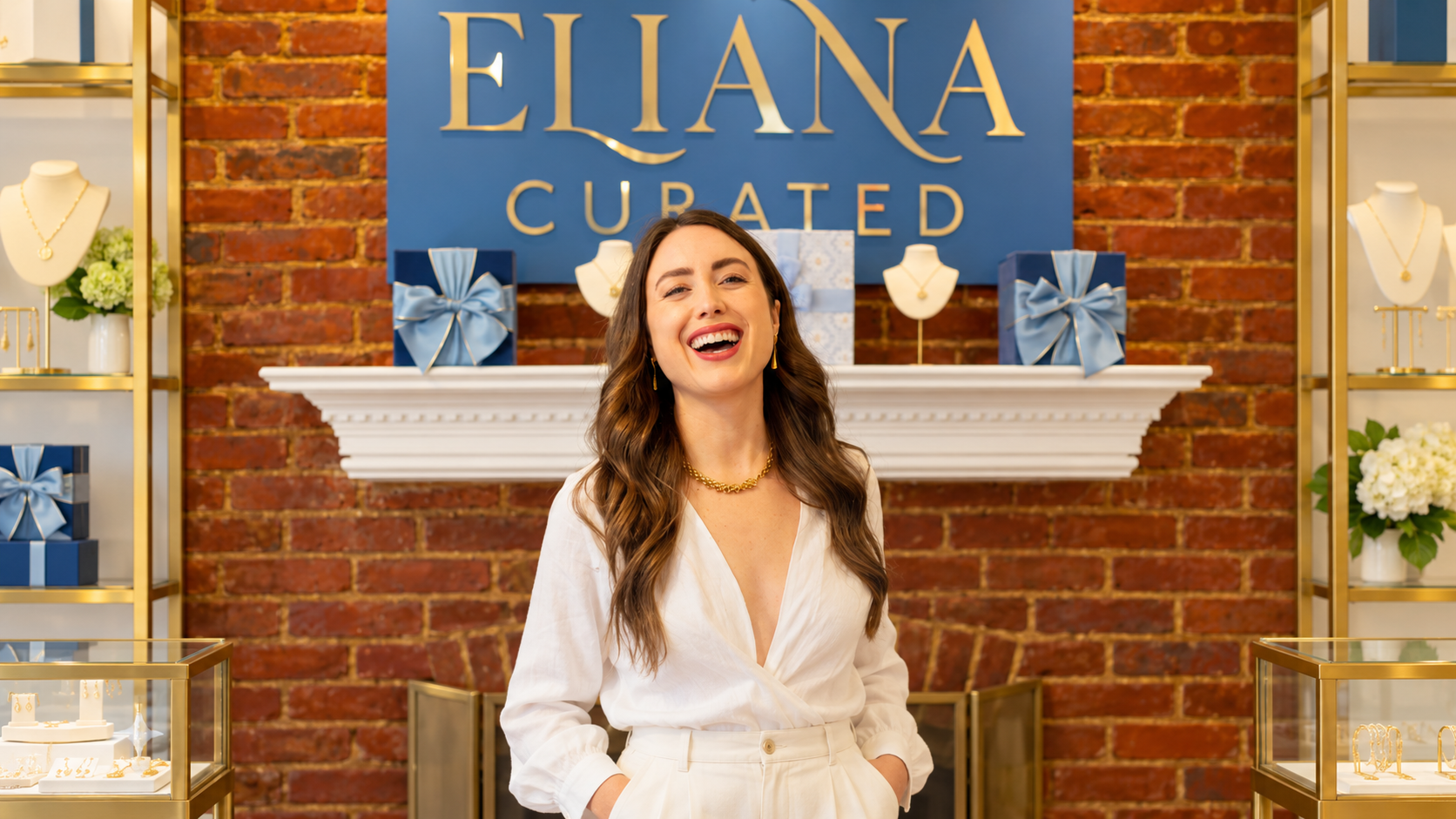 A smiling woman models gold jewelry inside Eliana Curated, with necklace displays and gift boxes beh 