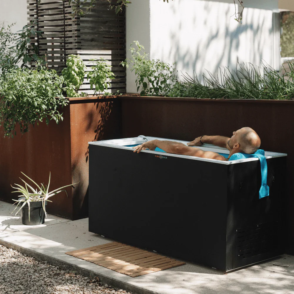 A bald man is relaxing in a black cold plunge tub outdoors. He is leaning back with his arms resting on the edge of the tub and his head supported by a blue towel. The tub is surrounded by greenery and a modern outdoor planter. A wooden mat is placed in front of the tub on the concrete ground.