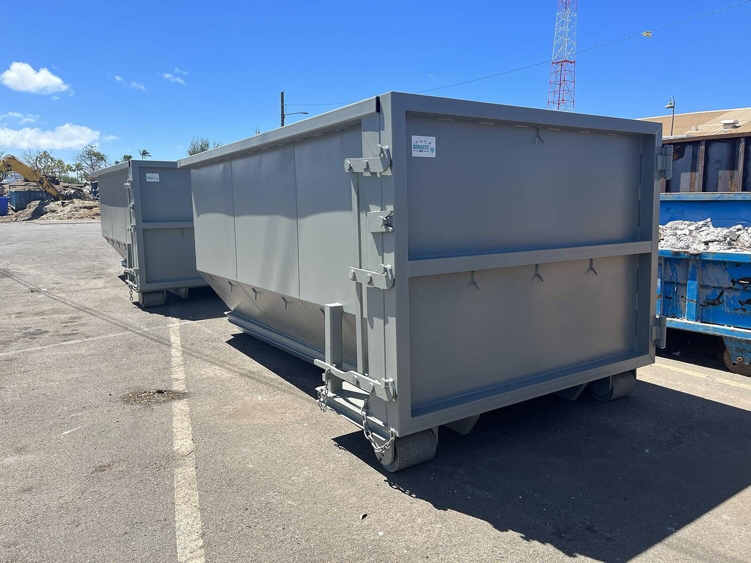 Large gray roll off dumpsters are lined up in an industrial yard under clear skies. The containers are used for construction debris, demolition waste, and large scale cleanup projects.