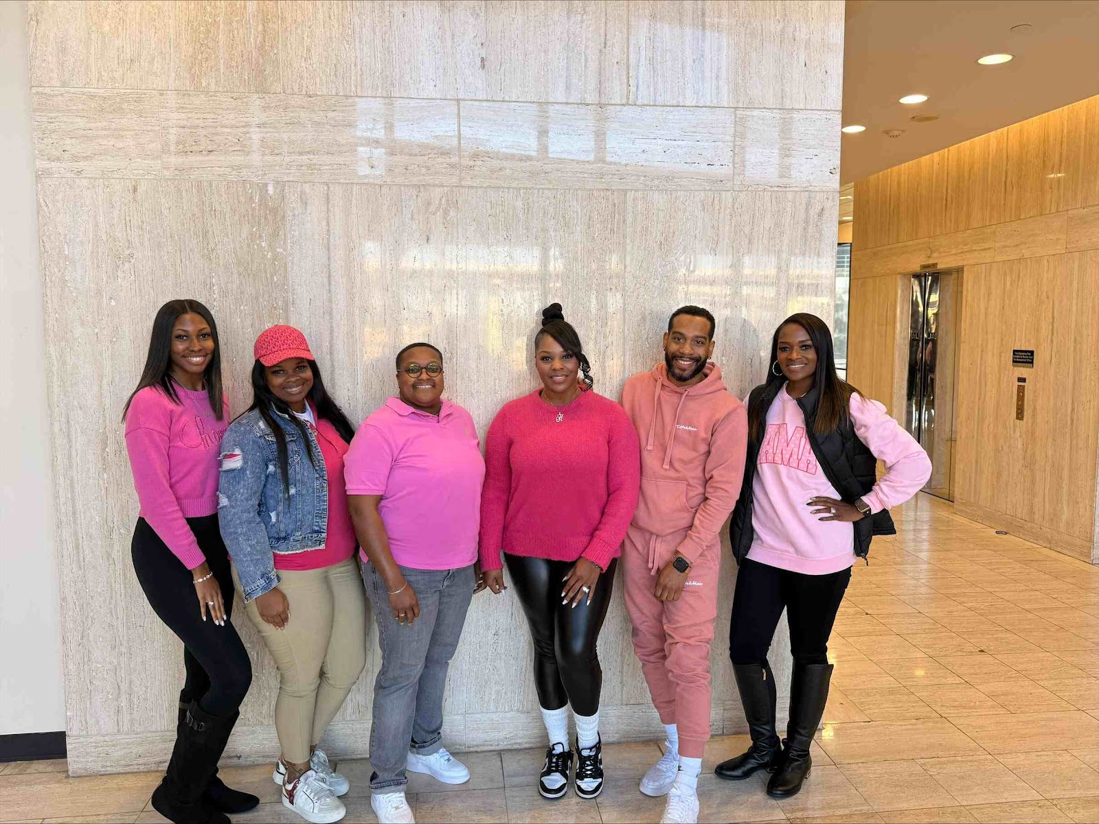 A group of six Black individuals, consisting of five women and one man, are posing for a photo in what appears to be a modern building lobby. All individuals are dressed in varying shades of pink, suggesting a coordinated theme. The background features a large, textured marble wall and a tiled floor, with an elevator visible to the right.