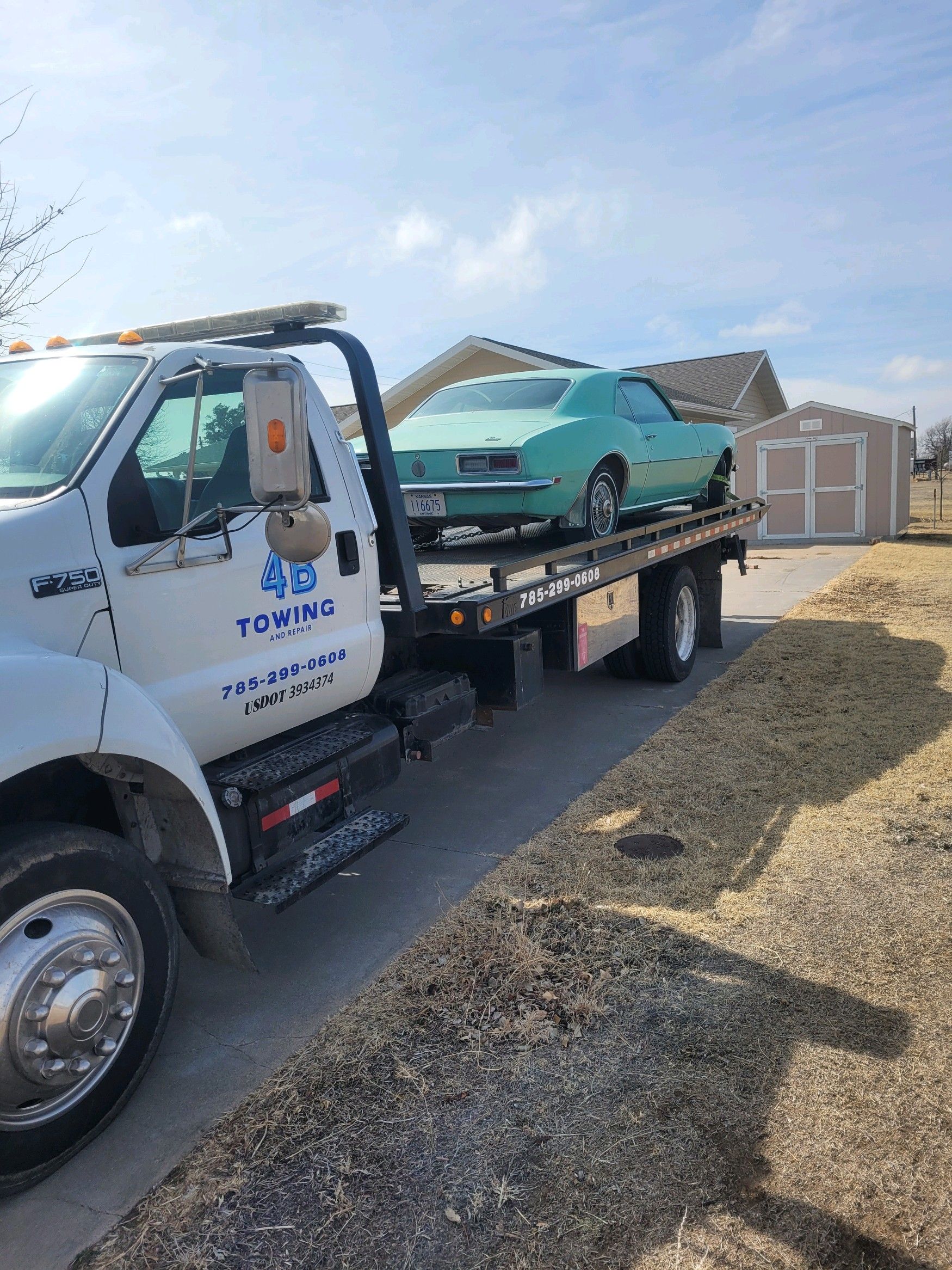 A light turquoise vintage classic car is loaded onto the back of a white 4B Towing flatbed truck parked in a residential driveway near a shed on a sunny day.