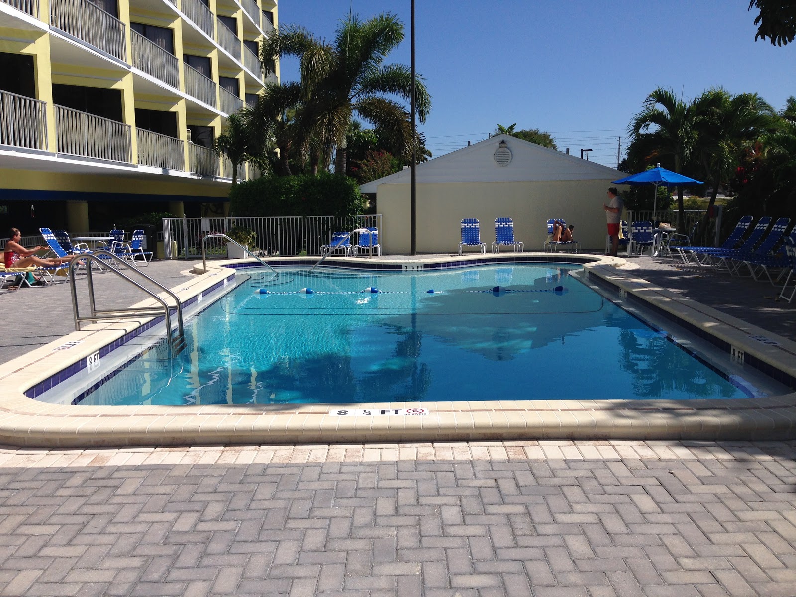 Commercial or condominium swimming pool with clear blue water, palm trees, lounge seating, and surrounding residential buildings.